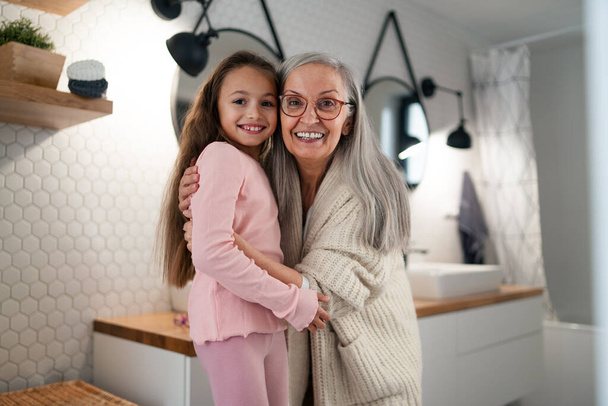 Senior grootmoeder en kleindochter staan binnen in de badkamer, knuffelen en kijken naar de camera. - Foto, afbeelding