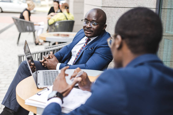 Two dark-skinned African American businessmen in suits and glasses with briefcases sit at a table in an outdoor city cafe. - 写真・画像