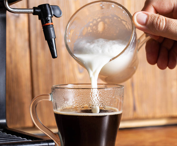 Nalít mléko do šálku s černou kávou na pozadí dřevěné desky stolu. Detailní záběr, kávovar s manuální cappuccino maker, aroma - Fotografie, Obrázek