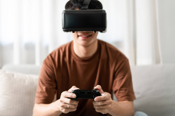 Conceito de tecnologia Uma pessoa usando um fone de ouvido de realidade virtual e segurando um jogo de console preto enquanto está sentado no sofá. - Foto, Imagem