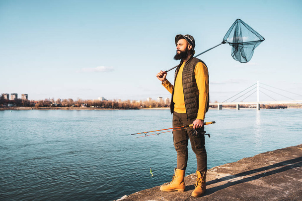 Image of fisherman holding fishing rod  and fishing net while standing by a river. - Valokuva, kuva
