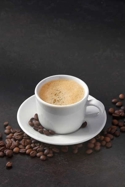 white cup of coffee on a saucer with coffee beans on a dark background. vertical image - Foto, Bild