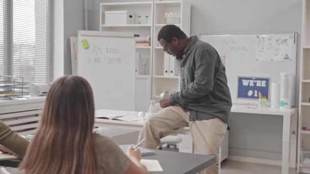 アフリカ系アメリカ人の男性教師が教室で子供のための化学のレッスンを行い、机に座ってコピー本でメモを取る生徒にプラスチック分子モデルを示します。 - 映像、動画