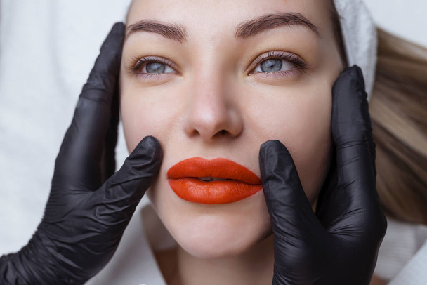 Постоянный макияж. Татуировка губ. Ярко-красные сексуальные губы в руках косметолога в черных перчатках - Фото, изображение
