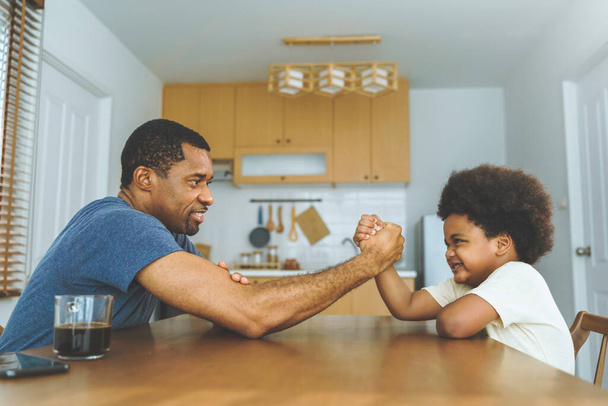 Zwarte Afro-Amerikaanse vader concurreren in arm worstelen met zijn kleine jongen in de keuken thuis - Foto, afbeelding
