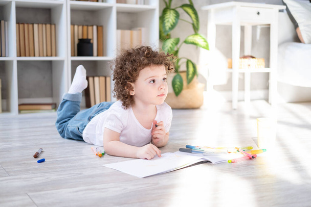 krullend kind meisje tekening met gekleurde markers liggen op de vloer in de woonkamer thuis - Foto, afbeelding