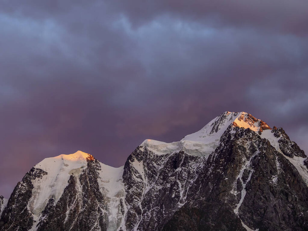 鋭い山々のシルエットの上に夜に赤い燃えるような雲。夕方の黄金の太陽と白い雪の点のピークでトップの黒い岩。山の中の壮大な紫の夕日.  - 写真・画像
