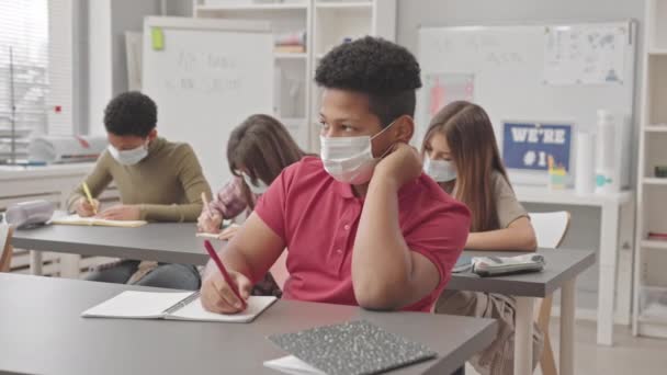 Περιμένετε μέχρι slowmo των 12 ετών Biracial αγόρι σχολείο στο πρόσωπο μάσκα κρατώντας σημειώσεις σε copybook κάθεται στο γραφείο με συμμαθητές στο μάθημα της επιστήμης - Πλάνα, βίντεο