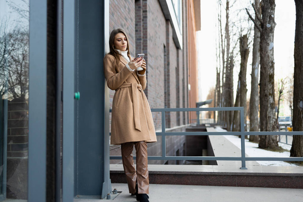 Beautiful young woman stands near the building with a cup of coffee and a smartphone - Fotó, kép