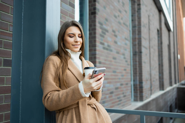 Young woman outdoors with a cup of coffee using smartphon - Фото, изображение