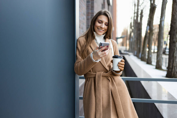 Linda chica morena en abrigo marrón con taza de café usando teléfono inteligente al aire libre - Foto, imagen