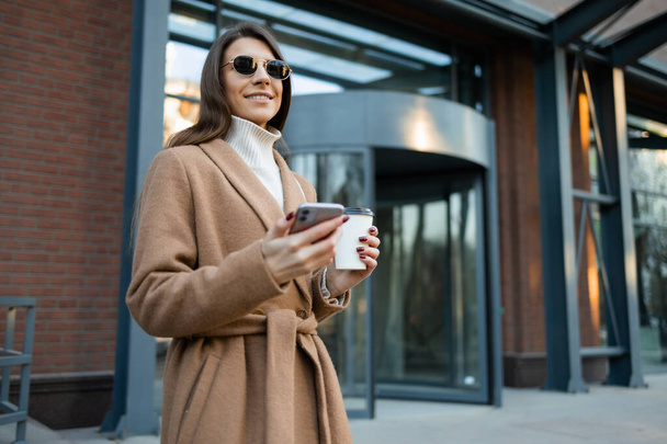 Stylish woman in a brown coat and glasses with a cup of coffee and a smartphone in her hands - Valokuva, kuva