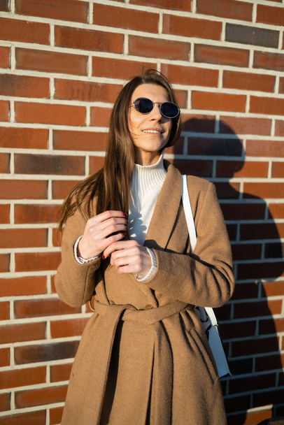 Stylowa młoda kobieta w brązowym płaszczu i okularach przeciwsłonecznych na tle cegły - Zdjęcie, obraz