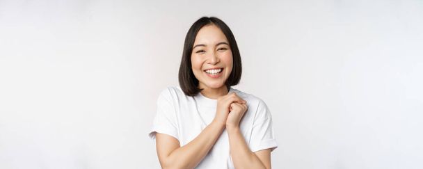 Retrato de una hermosa mujer coreana con una sonrisa blanca saludable, riendo y mirando feliz a la cámara, de pie en camiseta sobre fondo blanco del estudio - Foto, imagen