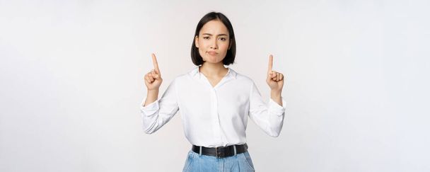 Kuva skeptinen aasialainen nainen irvistys, murjottaa järkyttynyt, osoittaa sormella ylös, näyttää mainos, seisoo valkoisella taustalla - Valokuva, kuva