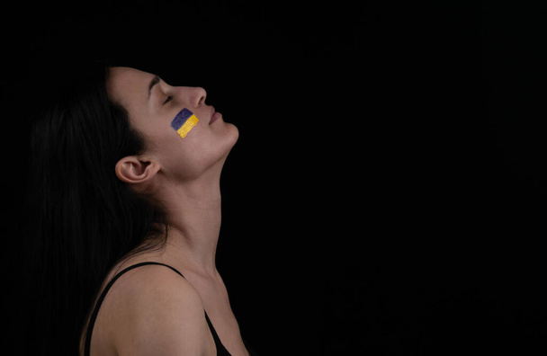 Πορτρέτο της όμορφης ενήλικης Ουκρανίας γυναίκα με κίτρινη μπλε σημαία στο πρόσωπό της. Πατριωτισμός. Ουκρανή καστανή κοπέλα κοιτάζει πάνω σε σκούρο φόντο. Χώρος για το κείμενό σας. - Φωτογραφία, εικόνα