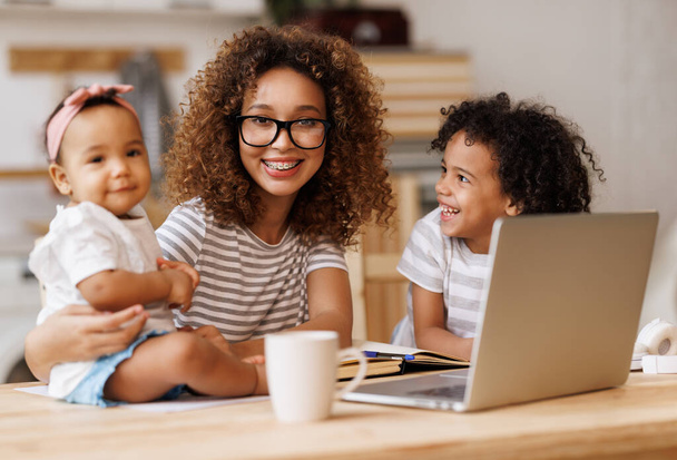 Young happy african american woman freelancer working remotely from home with two little kids - Photo, Image