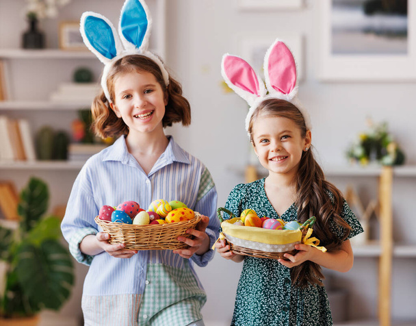 Дві позитивні маленькі дівчатка з вухами кролика посміхаються і тримають великодній кошик в руках вдома
 - Фото, зображення