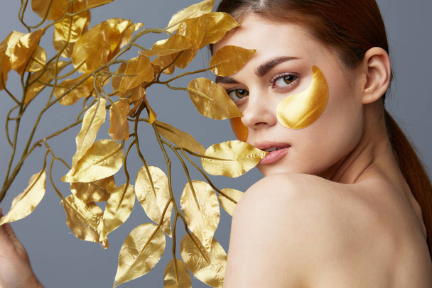 mujer joven de oro hojas de lujo cuidado de la piel parches para la cara hombros desnudos Fondo gris - Foto, imagen