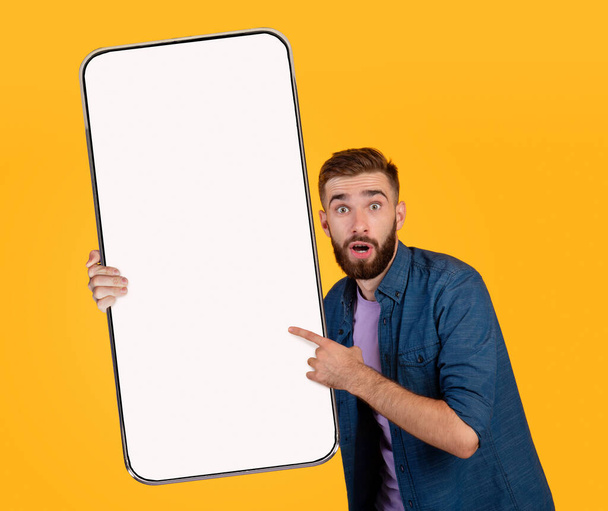 Shocking Offer. Excited young man holding and pointing at big blank smartphone - Photo, Image
