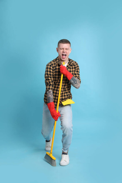 Handsome young man with floor brush singing on light blue background - Фото, изображение