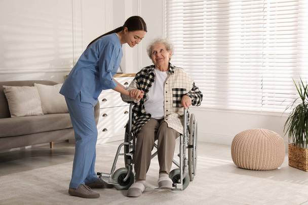 Young caregiver assisting senior woman in wheelchair indoors. Home health care service - Foto, immagini