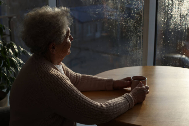 Elderly woman with drink looking out of window indoors on rainy day, space for text. Loneliness concept - Фото, изображение