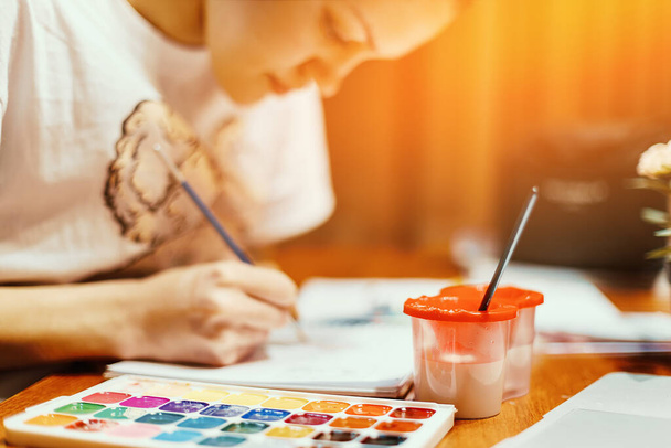 少女は部屋のテーブルに座り、水彩絵具で絵を描く。創造性、インスピレーション、ドローイングのテーマ. - 写真・画像