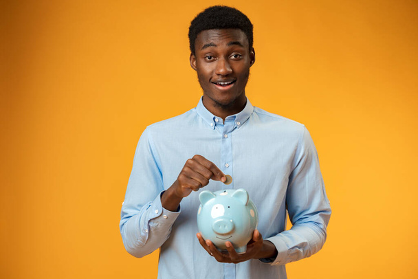 Young african amercian man holding piggy bank over yellow background - Foto, Bild