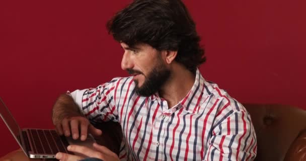 Fiatal üzletember dolgozik otthon laptoppal egy barna karosszéken, piros festett fallal a háttérben. Szürke jegyzetfüzet a munkához. Otthoni iroda koncepció. Kiváló minőségű videó - Felvétel, videó