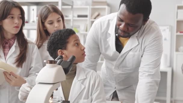 Mediana lentitud de la alumna afroamericana en bata de laboratorio usando microscopio en la clase de Ciencias mientras sus compañeras de clase y maestra de pie cerca observando y tomando notas - Imágenes, Vídeo