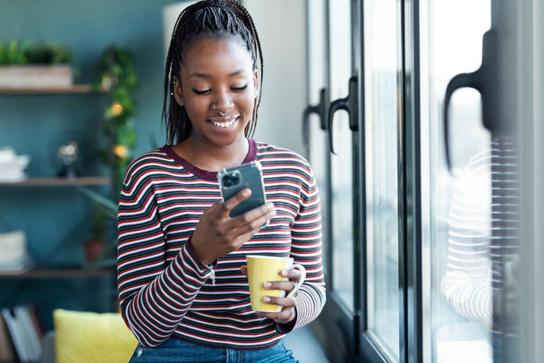 Shot of smiling young african american woman using her mobile phone while drinking a cup of coffee sitting in the sofa at home. - Valokuva, kuva