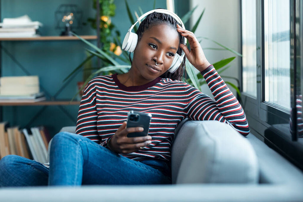 Shot van mooie vrouw luisteren naar muziek tijdens het gebruik van smartphone zitten op de bank thuis. - Foto, afbeelding