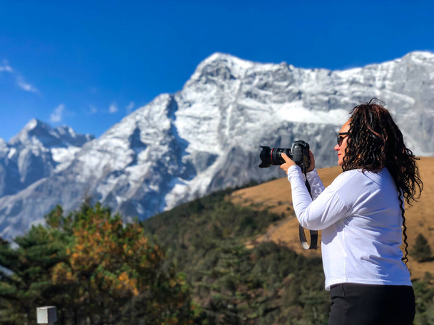 Γυναίκα με σγουρά μαλλιά γυρίσματα στην κάμερα του όμορφου πανοράματος του χιονισμένου βουνού Jade Dragon Χιόνι με κίτρινα λιβάδια και δάσος. Βρίσκεται κοντά στο Shangri - La. Θιβέτ, Κίνα - Φωτογραφία, εικόνα