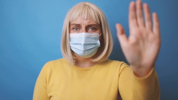 Porträt einer jungen Frau der 30er Jahre in einer medizinischen Schutzmaske mit gelbem Pullover, die eine Stop-Geste mit der Hand vor isoliertem blauen Hintergrund macht - Filmmaterial, Video