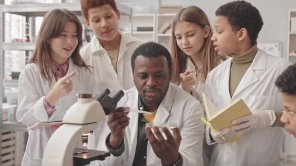 Plan moyen d'élèves du collège multiethnique et de leur professeur afro-américain en blouse de laboratoire regardant des bactéries sur une boîte de Pétri au microscope en classe de sciences - Séquence, vidéo