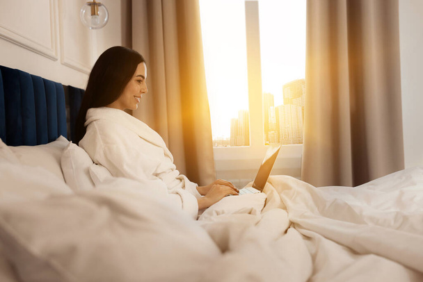 Happy young woman working with laptop on bed in hotel room - Photo, Image