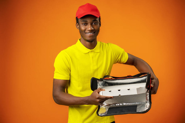 Smiling black male courier wearing uniform holding box in orange studio - Fotografie, Obrázek
