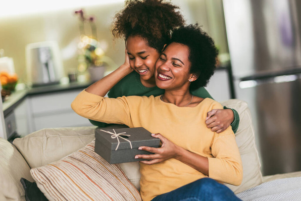 Mujer y niña en casa celebrando el día de la madre sentadas en el sofá hija sosteniendo caja de regalo para la madre. - Foto, imagen