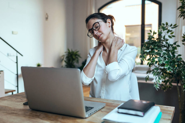 Shot of tired business woman with neck pain looking uncomfortable while working with laptop in the office. - Foto, afbeelding