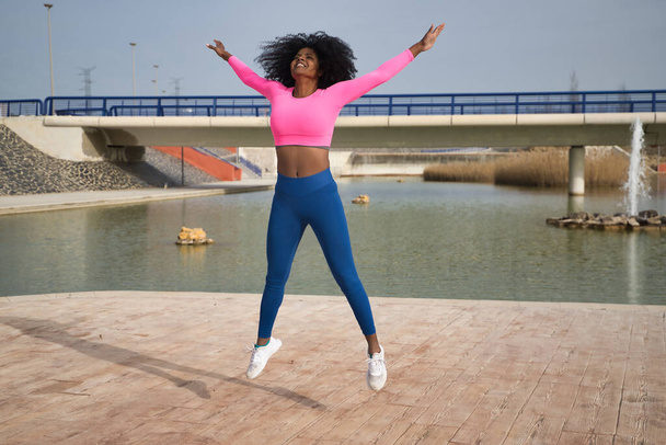 Donna afro-americana con capelli afro e abbigliamento sportivo, con t-shirt rosa fluo e leggings, facendo esercizi di salto accanto a un lago all'aperto. Fitness concept, sport, strada, urbano. - Foto, immagini