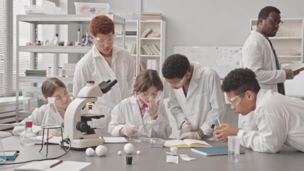 Lentidão média do grupo de alunos multiétnicos inteligentes em casacos de laboratório e óculos de proteção conduzindo experimento de laboratório com amostra em placa de Petri tendo aula de ciência em sala de aula moderna - Filmagem, Vídeo