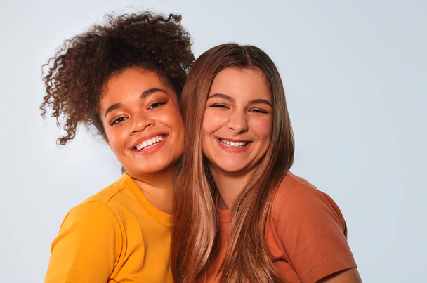 Positive multiracial teen girls, interracial best friends, standing close to each other, gently snuggling cheek to cheek, looking at camera smiling honestly and brightly against white wall background - Photo, Image