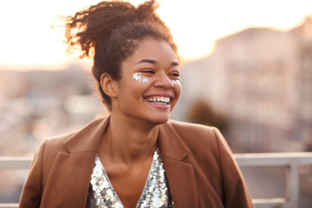 Retrato al aire libre de la mujer afroamericana glamour feliz con brillo en la cara con vestido de lentejuelas de desgaste festivo de pie en la terraza de la azotea durante la fiesta o la celebración y sonriendo felizmente  - Foto, imagen