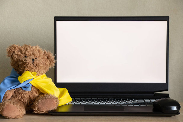 Teddy bear with flag of Ukraine near laptop with white screen on table at home, war in Ukraine, patriot, template, peace in Ukraine, screen with white screen - Foto, imagen