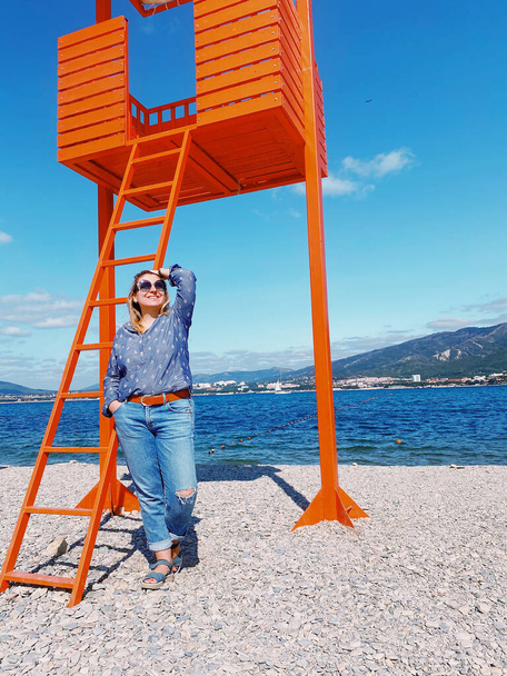 Glückliche junge Frau in legerer Kleidung steht am Strand an einem sonnigen Sommertag in der Nähe von hölzernen orangefarbenen Rettungsschwimmer stehen mit leuchtend blauem Meerwasser auf dem Hintergrund, genießen Urlaub und Sommer am Meer - Foto, Bild