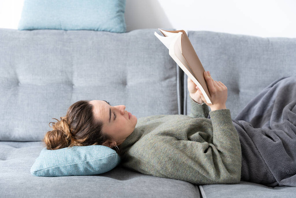 νεαρή γυναίκα ξαπλωμένη στον καναπέ και διαβάζοντας ένα βιβλίο - Φωτογραφία, εικόνα