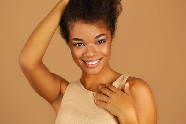 Close up portret mieszanej rasy młoda dziewczyna patrząc na aparat z uśmiechniętym wyrazem twarzy, trzymając się za ręce blisko szyi stojąc izolowany nad beżowym tle ściany studio - Zdjęcie, obraz