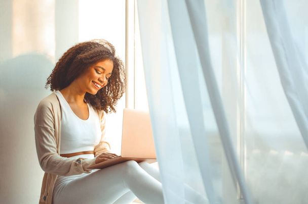 Cheerful afro american young lady with curly hair sits on windowsill with laptop computer on laps, typing on keyboard looking at screen and smiling, happy african woman chatting with friends online - Fotografie, Obrázek
