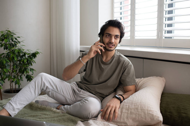Młody współczesny mężczyzna pracownik lub freelancer rozmawia przez telefon komórkowy siedząc na łóżku naprzeciwko okna - Zdjęcie, obraz